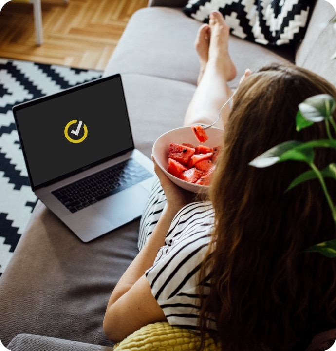 Mulher deitada em um sofá comendo uma tigela de frutas com um laptop. Exibindo ao lado dela o logotipo Norton